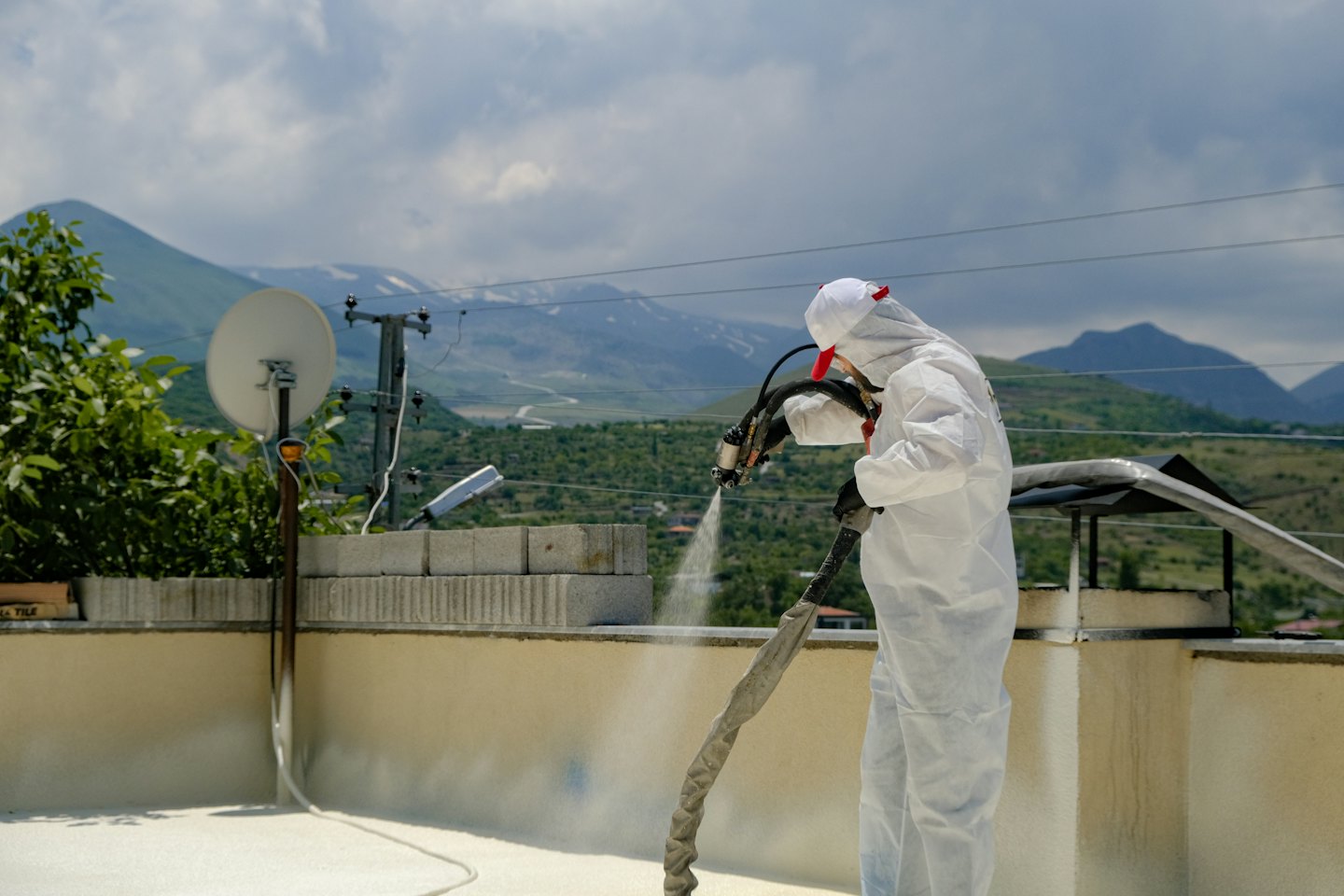 Un travailleur en combinaison de protection pulvérise de l'isolant sur un toit sous un ciel couvert.